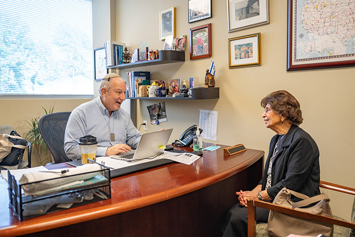 The image depicts a man sitting at a desk with a laptop, smiling and looking towards a woman who is standing in front of him, also smiling and engaging in conversation.