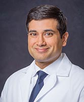 The image shows a man wearing a white lab coat, smiling slightly, with his head turned towards the camera. He has short dark hair, appears to be of South Asian descent, and is looking directly at the viewer.
