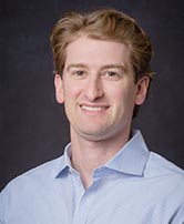 The image shows a man with short hair, wearing a light blue shirt, smiling at the camera against a dark background.