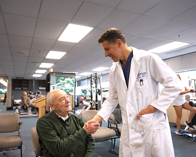 In the image, a man in a white lab coat is shaking hands with an older gentleman who appears to be a patient, both are seated in a gym setting, with exercise equipment visible behind them.