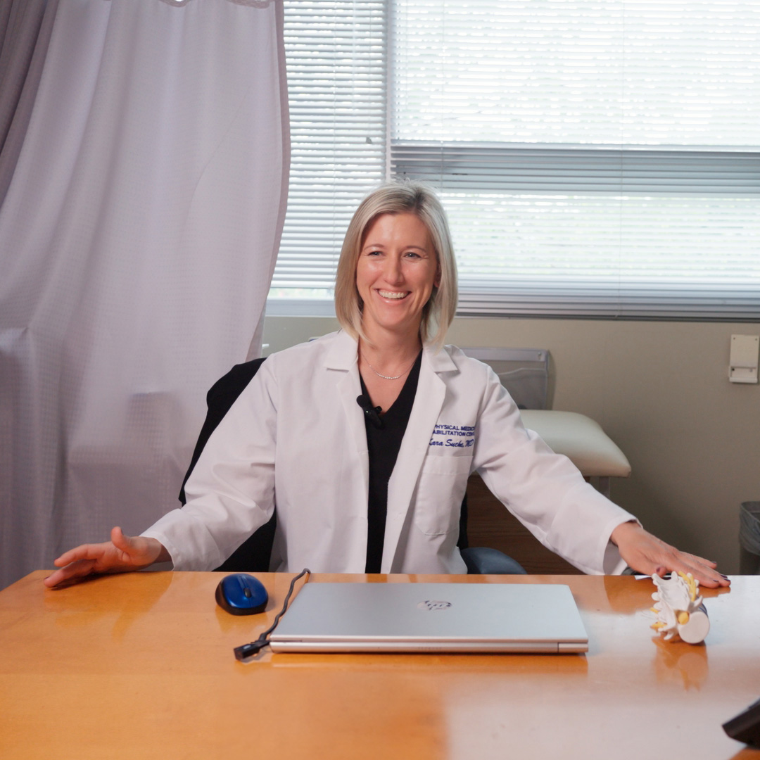 A female doctor sitting at a desk with her hands on her knees, smiling towards the camera.